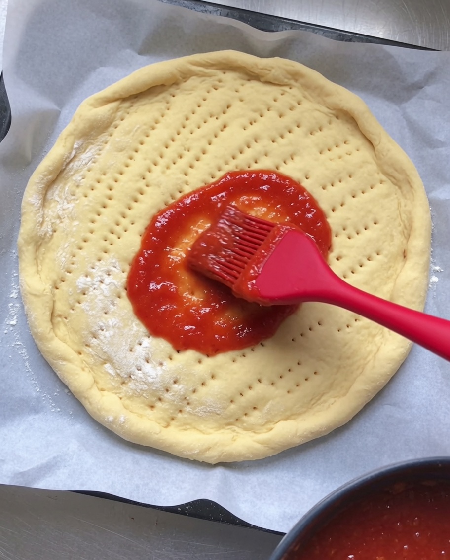 Rote Tomatensoße wird gleichmäßig auf einem runden, eingestochenen rohen Pizzateig verteilt.