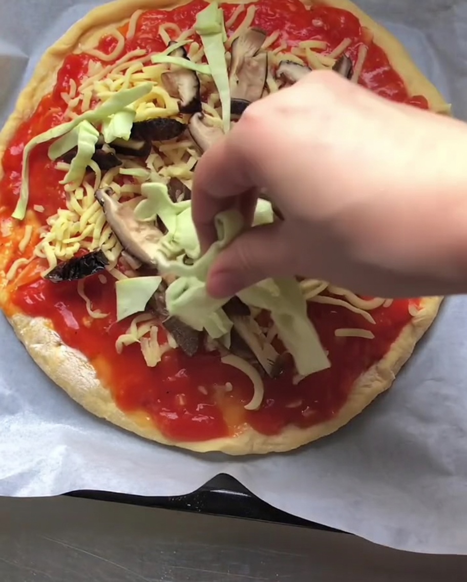 Eine Hand streut frischen, feingeschnittenen rohen Weißkohl über eine mit Tomatensoße, Käse und geschnittenen Pilzen belegte Pizza.
