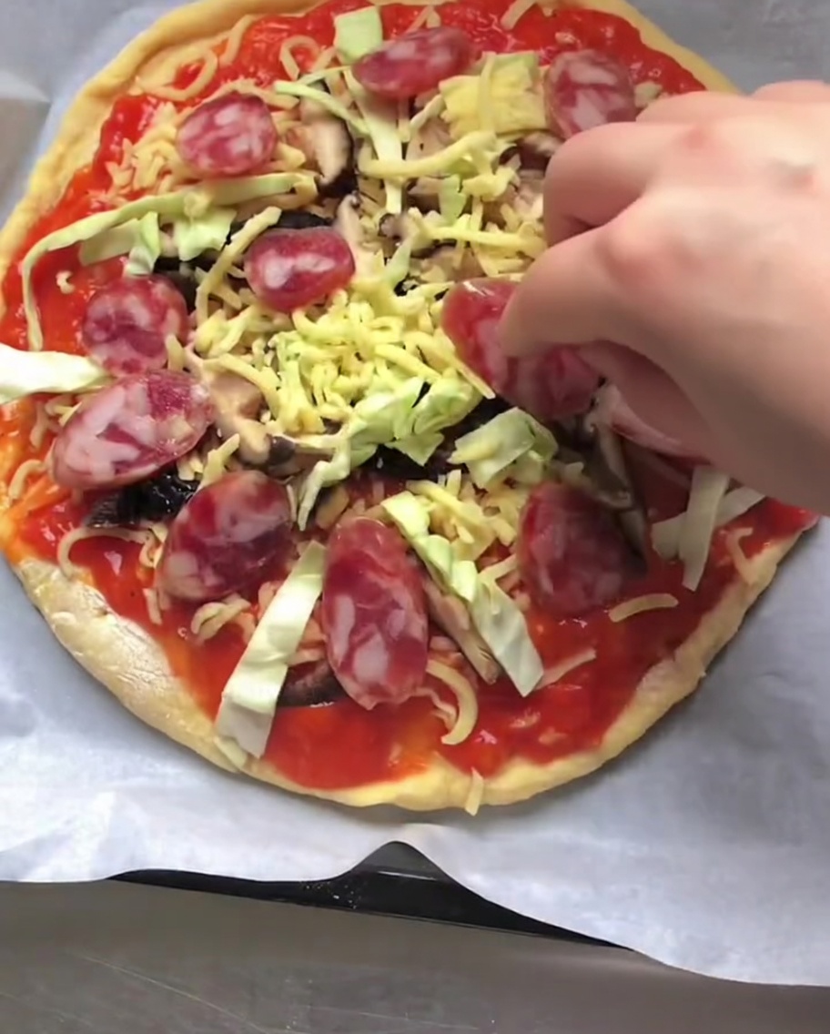 Eine Hand legt Scheiben von roter italienischer Salsiccia auf eine ungebackene Pizza mit Soße, Käse und Gemüse.