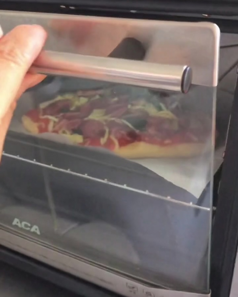 Die Tür eines kleinen Tischofens wird geöffnet, um eine rohe, belegte Pizza auf einem Backblech hineinzuschieben.
