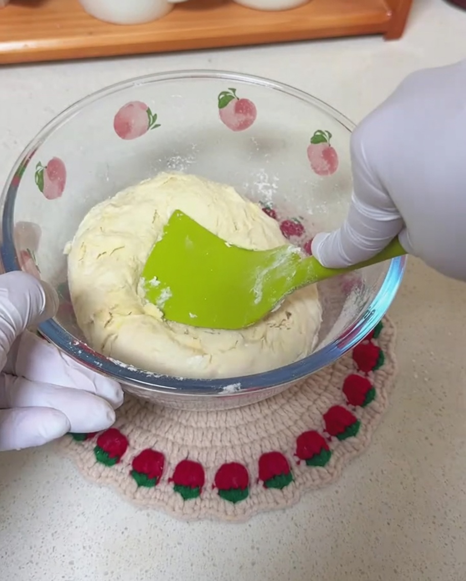 Hände mit Handschuhen mischen mit einem grünen Silikonspatel einen groben Teig in einer Glasschüssel.