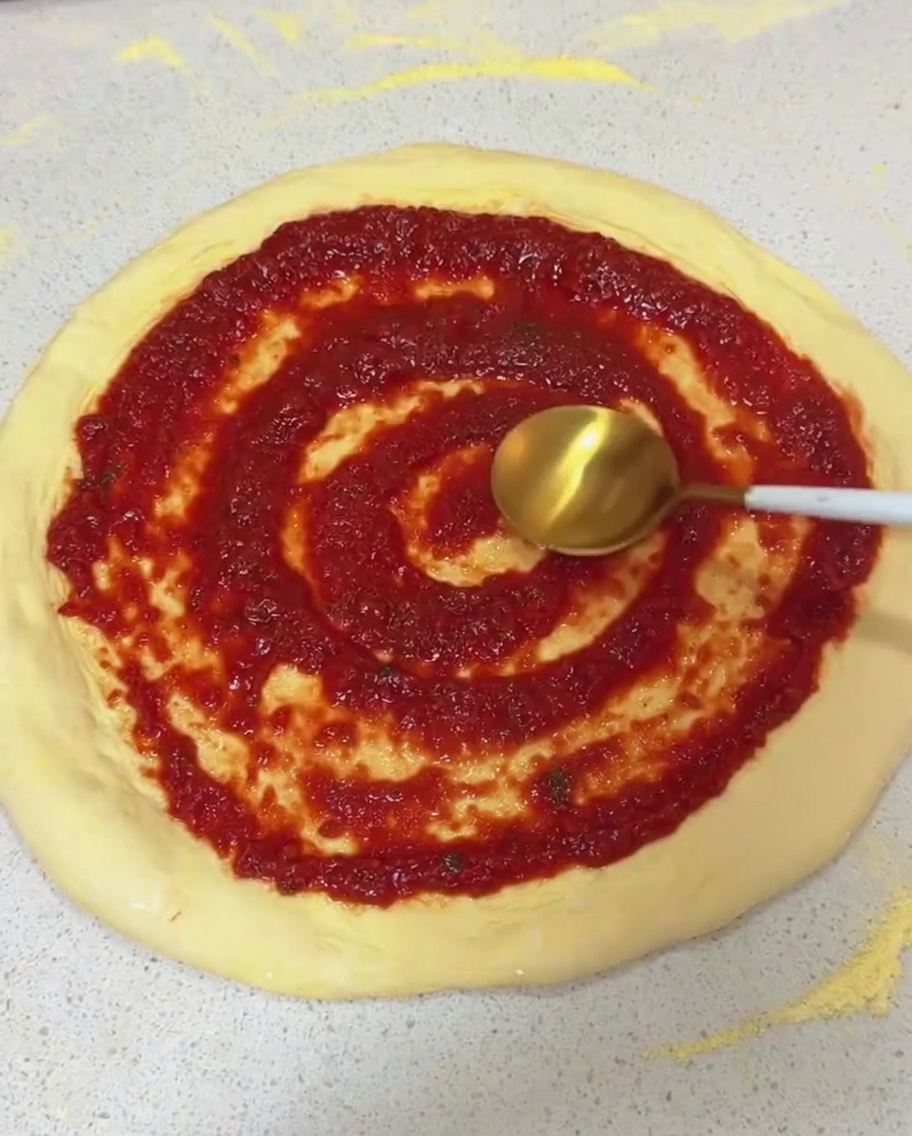 Verteilen der roten Tomatensauce in kreisenden Bewegungen auf dem rohen Pizzateig mit dem Rücken eines Löffels.