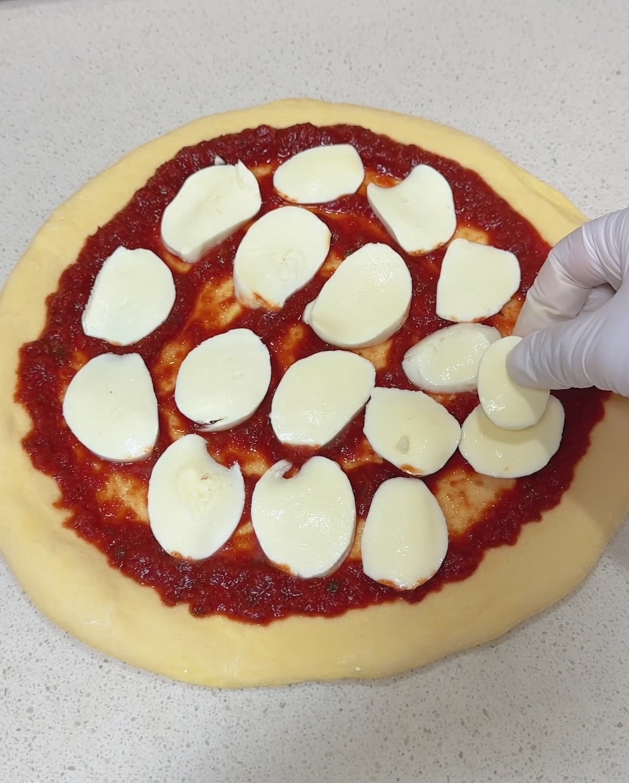 Eine Hand legt dicke Scheiben frischen Mozzarella auf einen mit Tomatensauce bestrichenen Pizzaboden.