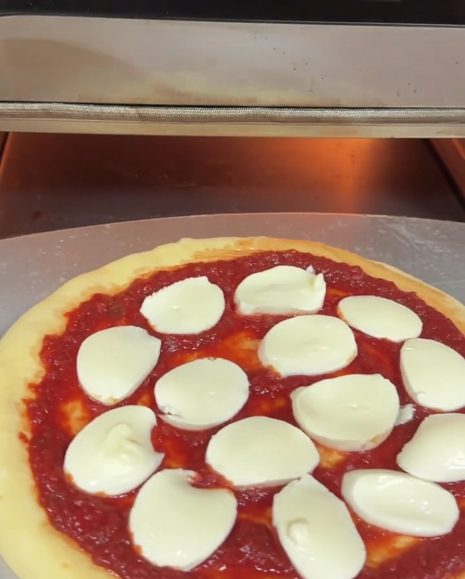 Eine ungebackene Pizza mit Sauce und Mozzarella wird auf einem Metallschieber in einen elektrischen Pizzaofen geschoben.