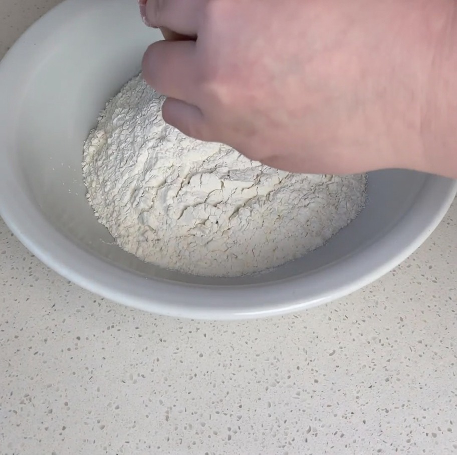 Hände schlagen ein rohes Ei in eine Mulde aus weißem, proteinreichem Mehl in einer Rührschüssel.