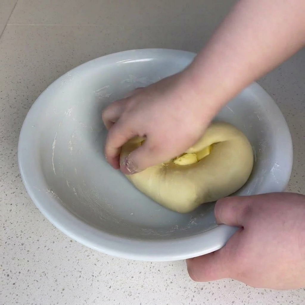 Hände kneten ein kleines Stück Butter in eine Teigkugel in einer großen weißen Schüssel.