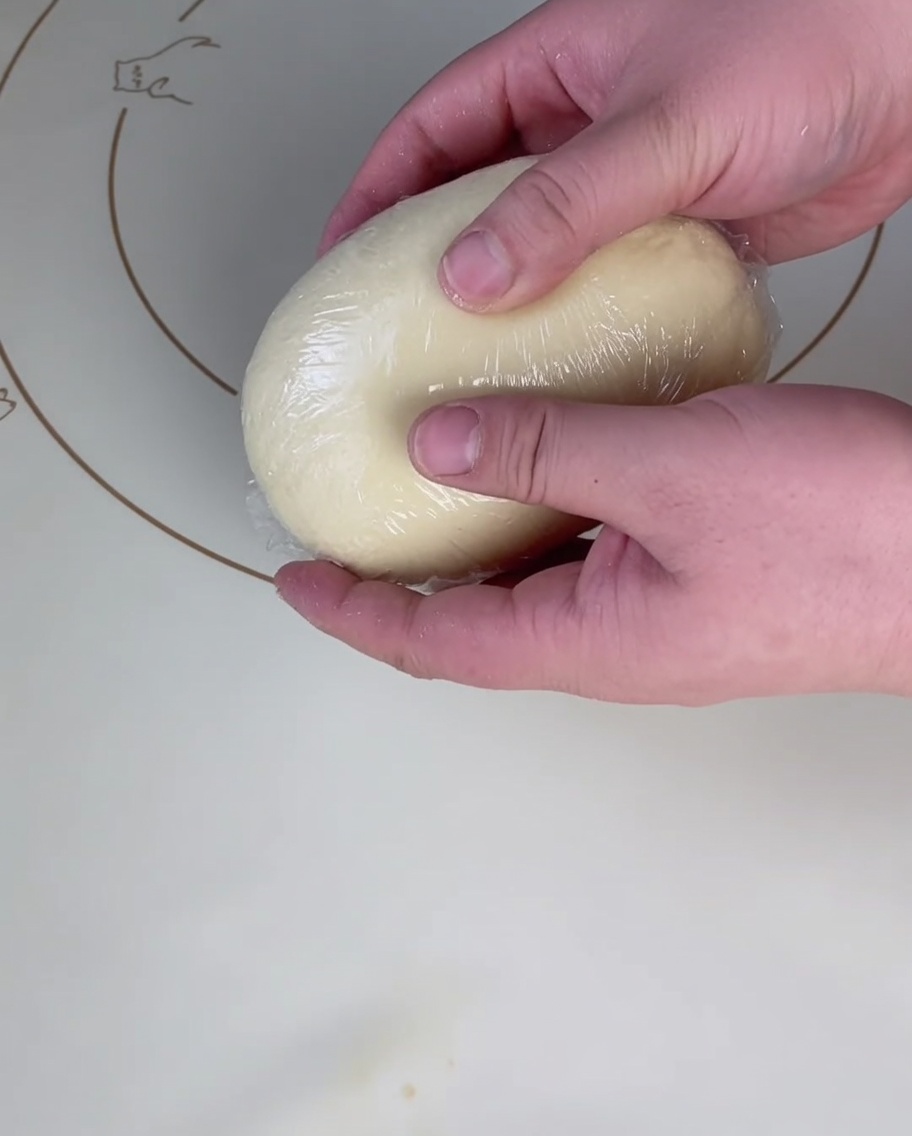 Eine glatte, runde Kugel Pizzateig wird auf einer Silikonbackmatte fest in Frischhaltefolie eingewickelt.