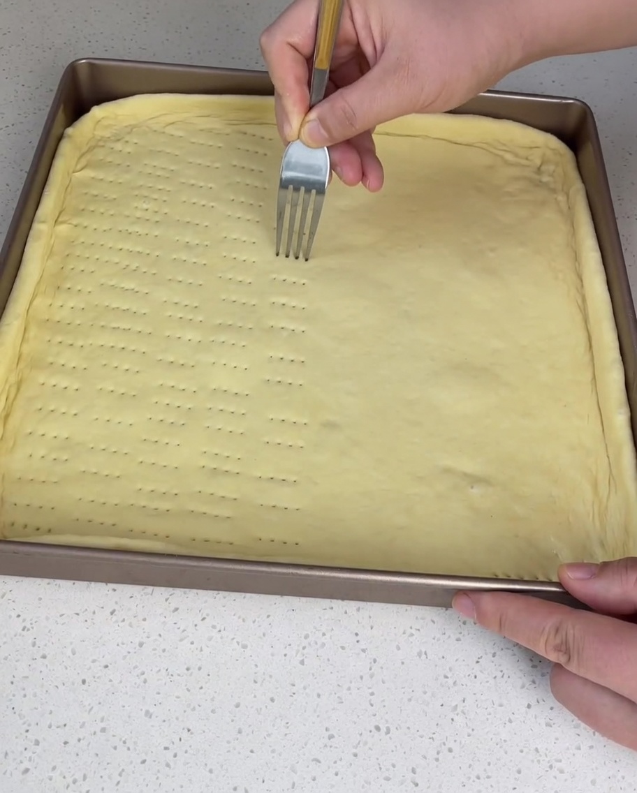 Mit einer Gabel werden mehrere Reihen kleiner Löcher in den rohen Pizzateig in einem quadratischen Backblech gestochen.