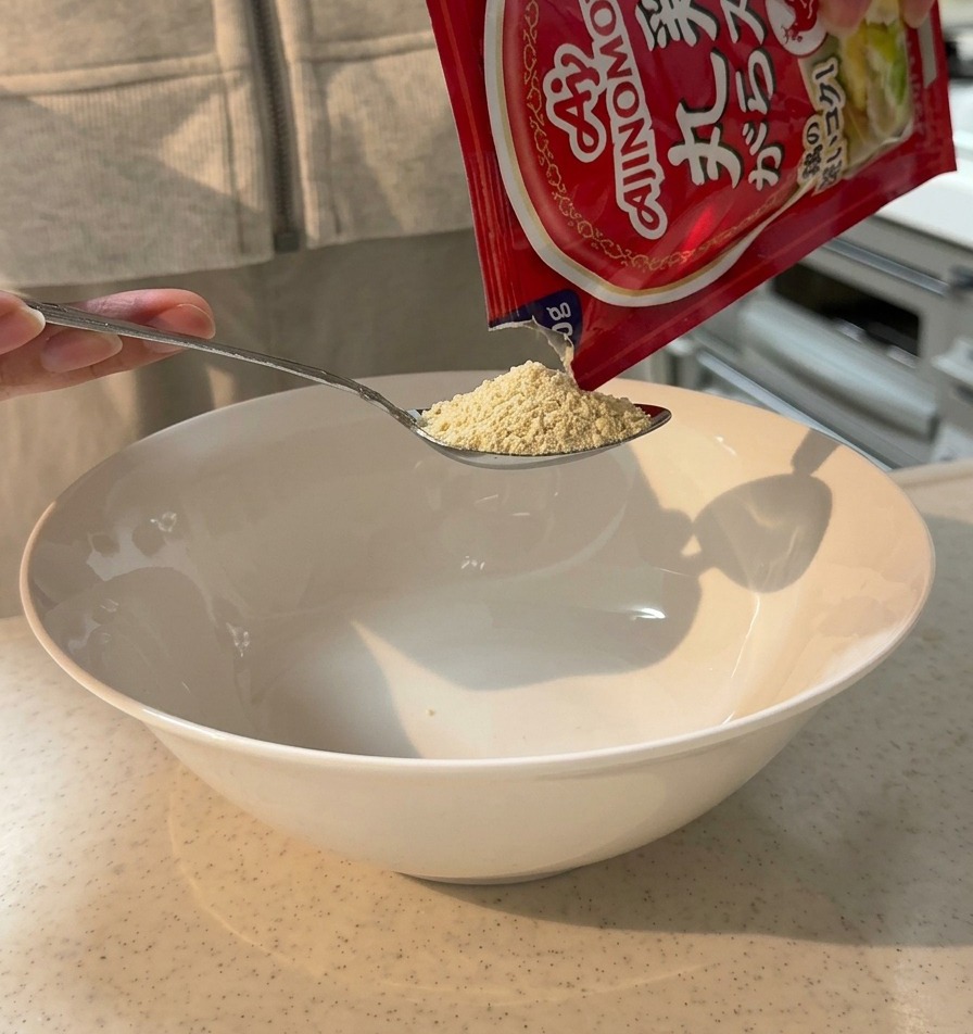 Hühnerbrühepulver wird aus einem Beutel mit einem Löffel in eine große weiße Servierschüssel gegeben.