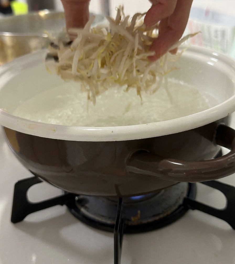 Eine Hand gibt frische Sojasprossen in einen Topf mit kochendem Wasser.