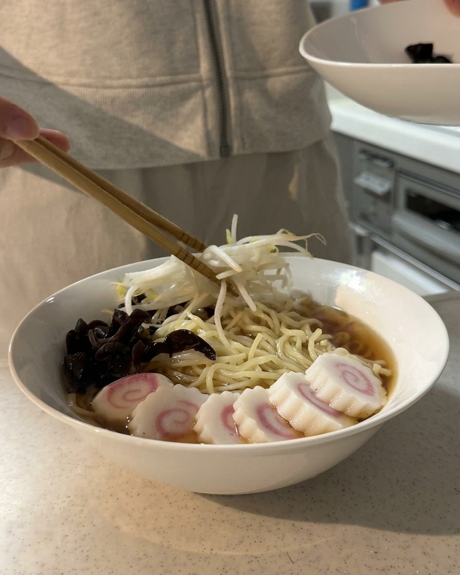 Stäbchen platzieren Sojasprossen auf einer Schüssel mit Ramen, die bereits mit Pilzen und Fischkuchen garniert ist.