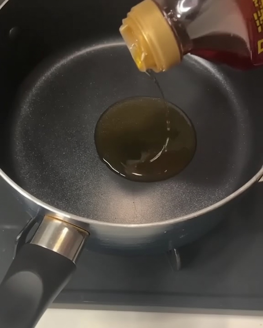 Eine Flasche, aus der klares, goldenes Sesamöl in einen leeren schwarzen Topf auf dem Herd gegossen wird.