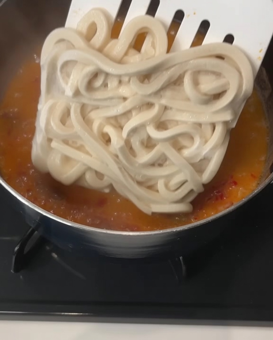 Ein großer, quadratischer Block dicker, gefrorener Udon-Nudeln wird mit einem Schaumlöffel in eine blubbernde, cremig-orangefarbene Brühe getaucht.