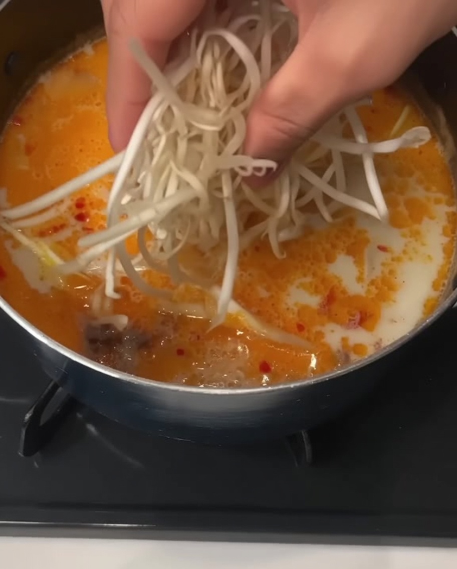 Eine Hand gibt eine Handvoll frischer weißer Sojasprossen in einen Topf mit cremiger Nudelsuppe.