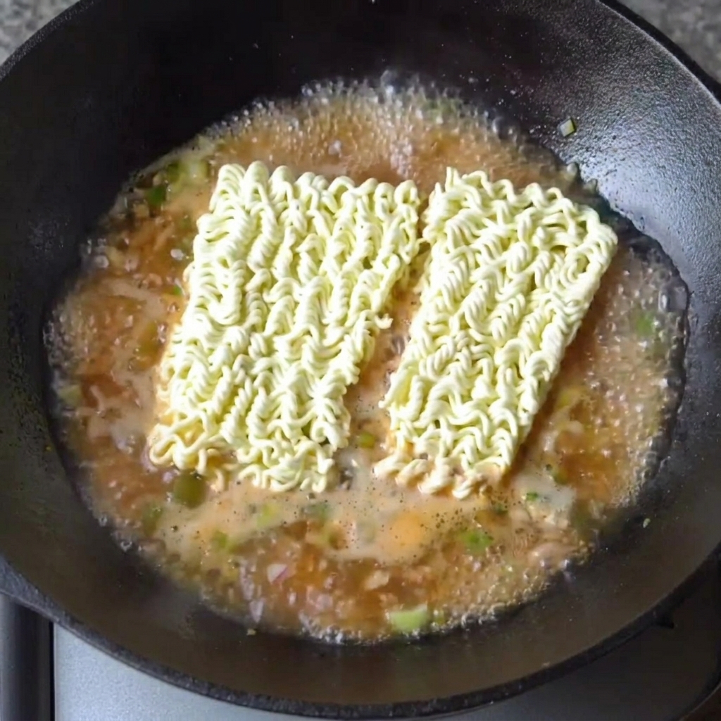 Zwei Blöcke getrocknete Ramen-Nudeln liegen in der siedenden, würzigen Brühe.
