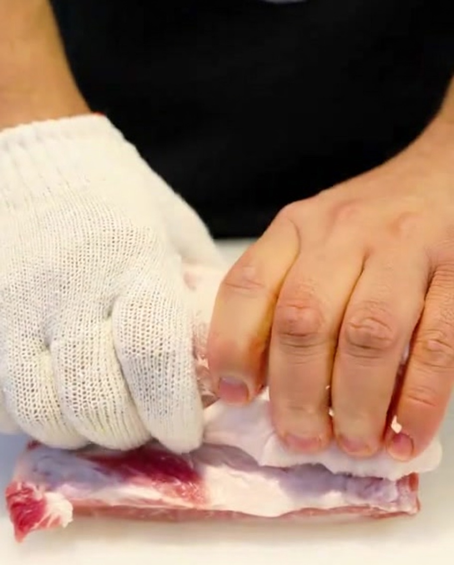 Eine rohe Schweinebauchrolle wird mit Küchengarn fest gebunden, die Hände tragen Schutzhandschuhe.