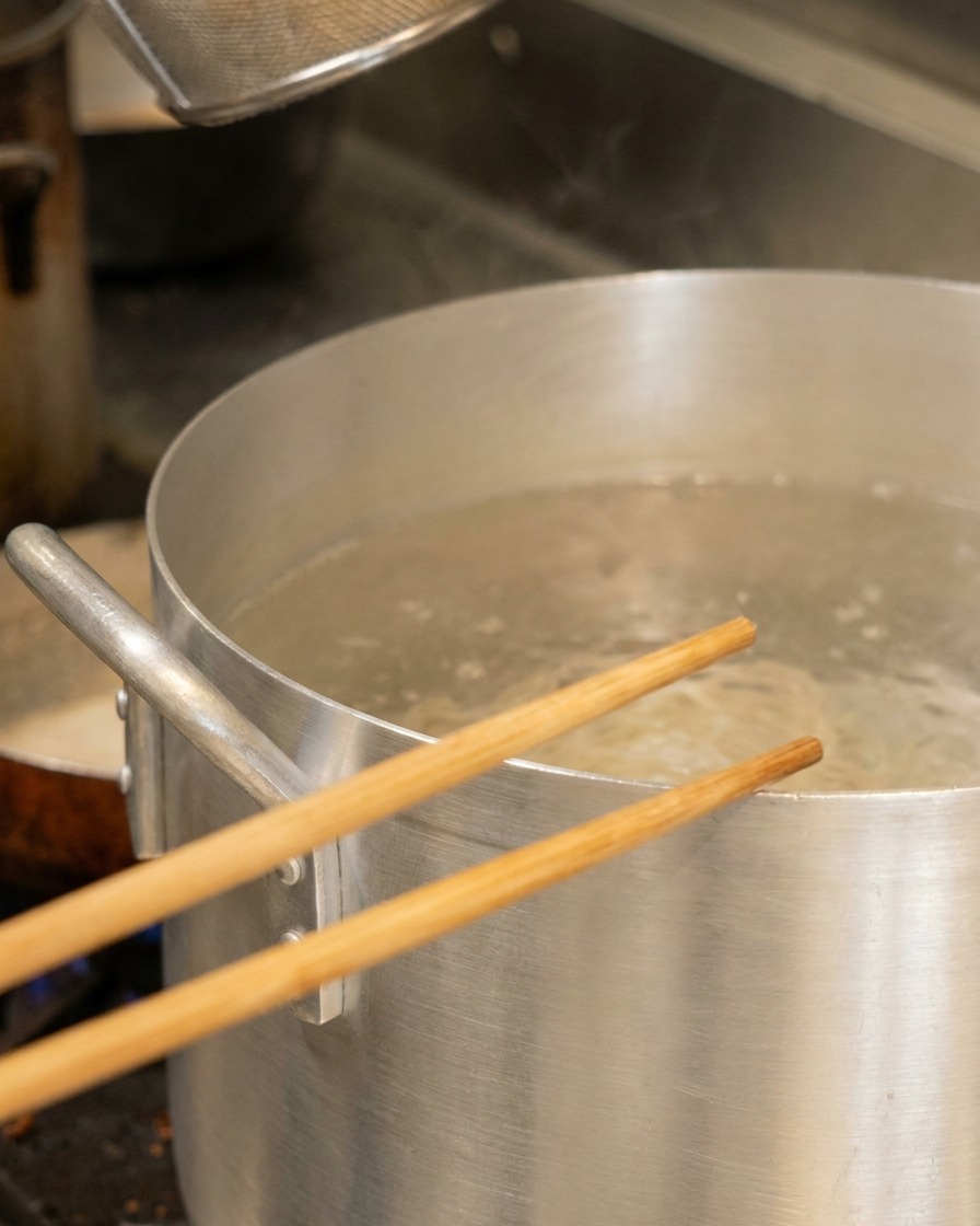 Holzstäbchen ruhen über einem großen Aluminiumtopf mit kochendem Wasser und einem Nudelsieb.