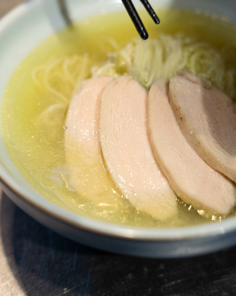 Essstäbchen legen zarte Hühner-Chashu-Scheiben über frisch zubereitete Ramen in einer Schüssel.