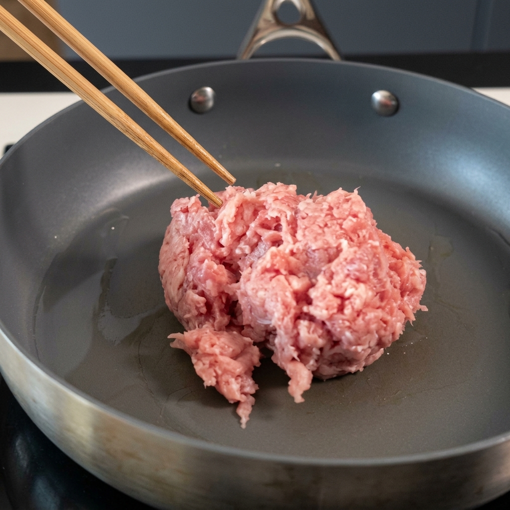 Schweinehackfleisch wird mit Stäbchen in einer beschichteten Pfanne angebraten.