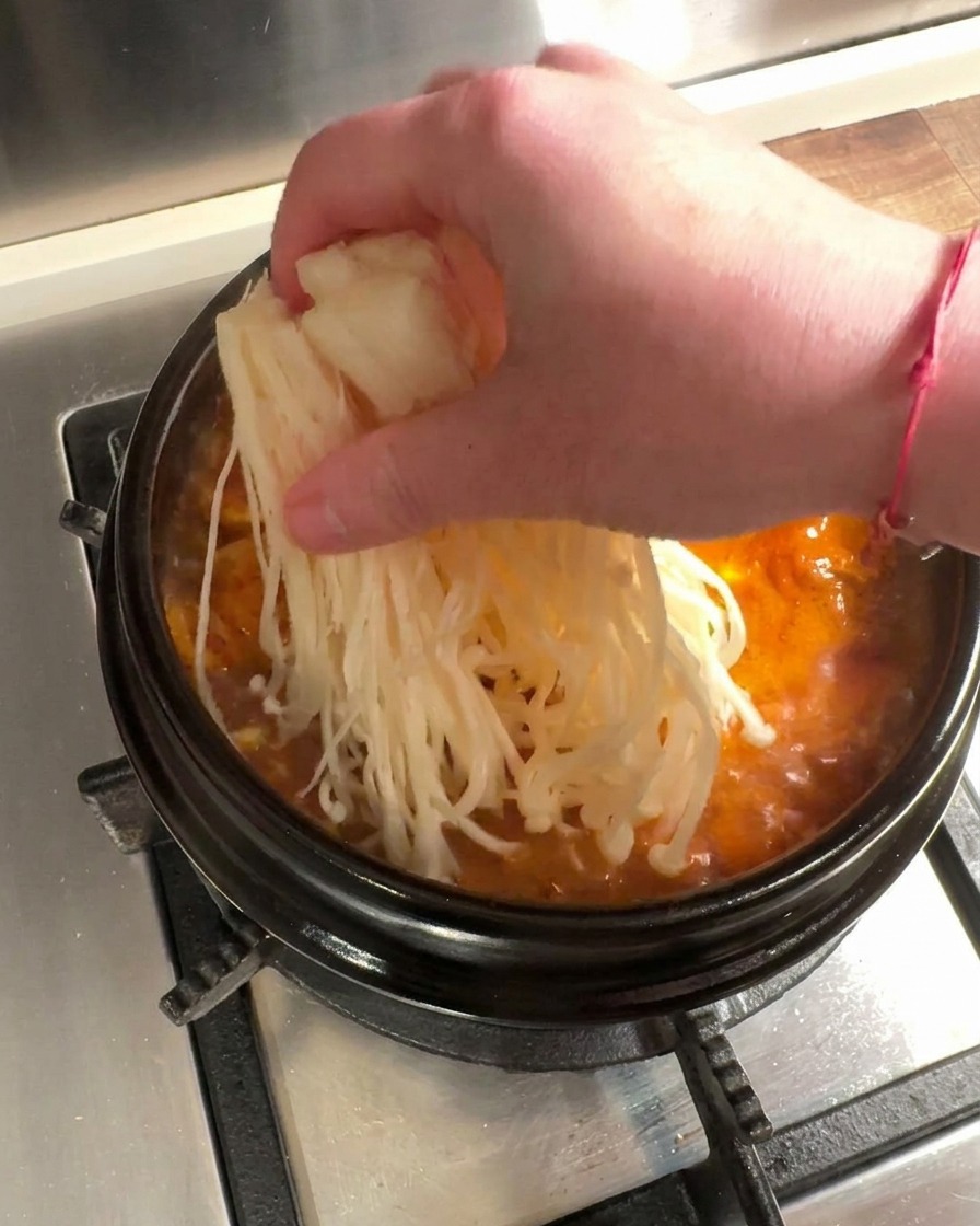 Frische Enoki-Pilze und in dünne Scheiben geschnittenes rohes Rindfleisch werden in einen brodelnden, roten Kimchi-Eintopf gelegt.