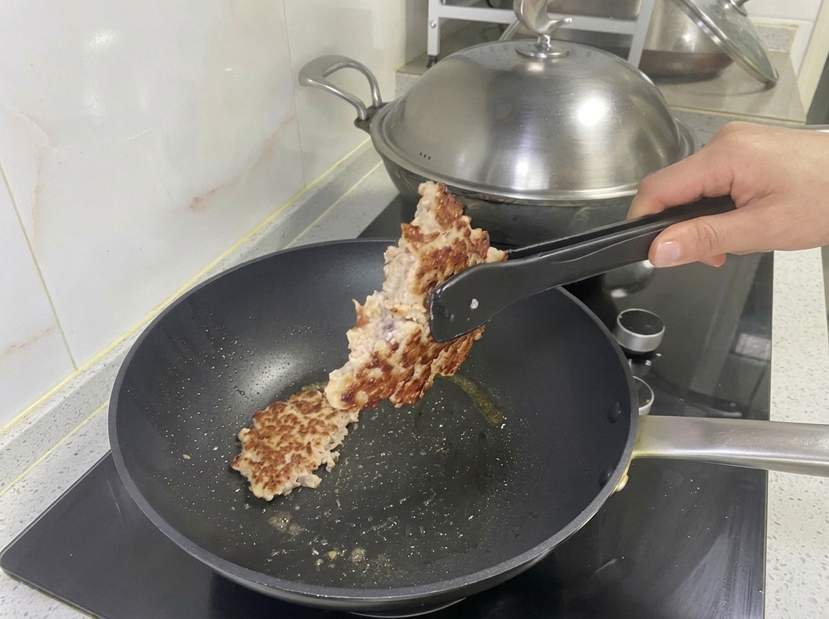 Ein gebratenes, goldbraunes Schweinefleisch-Patty wird mit einer Zange aus der Pfanne gehoben.