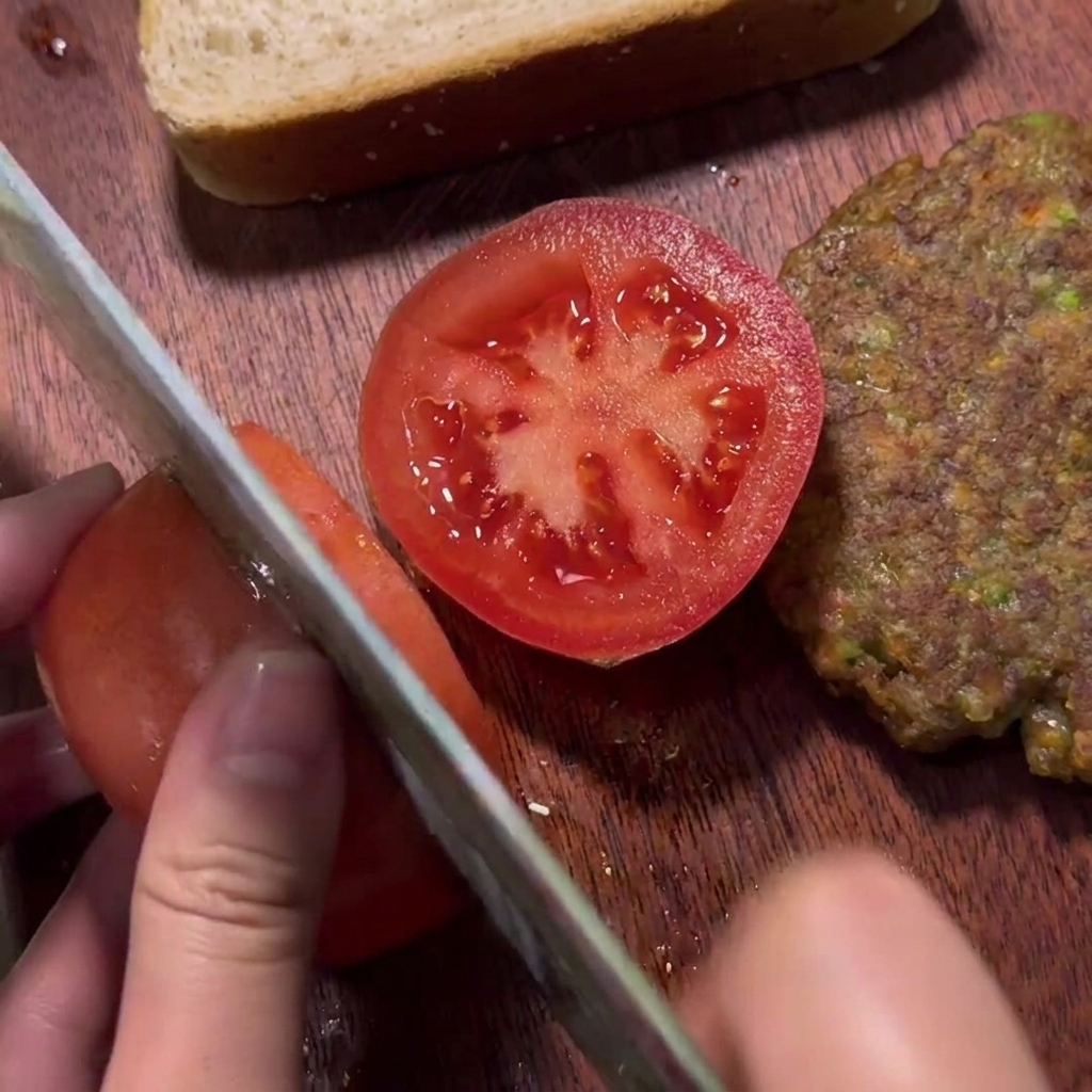 Eine Hand schneidet eine frische rote Tomate auf einem Holzbrett neben einem gebratenen Rinder-Patty.