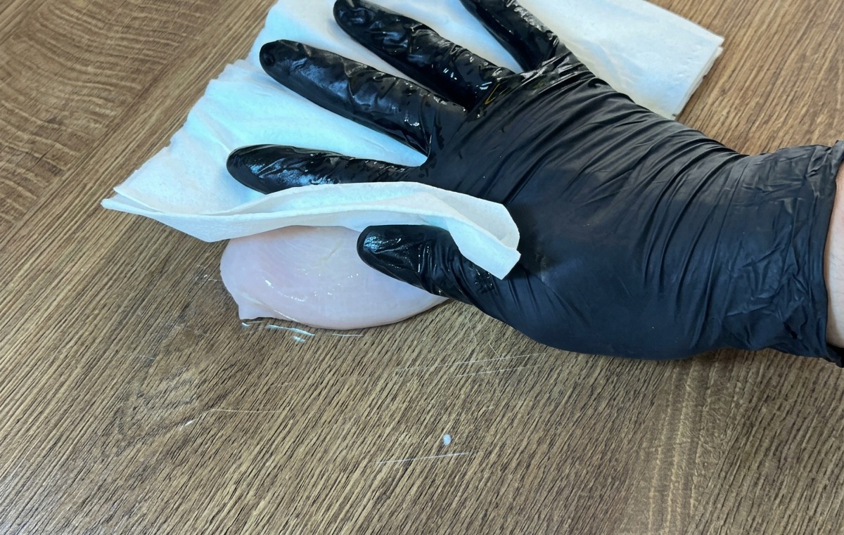 Ein behandschuhter Hand tupft die rohe Hähnchenbrust mit einem Papiertuch trocken.
