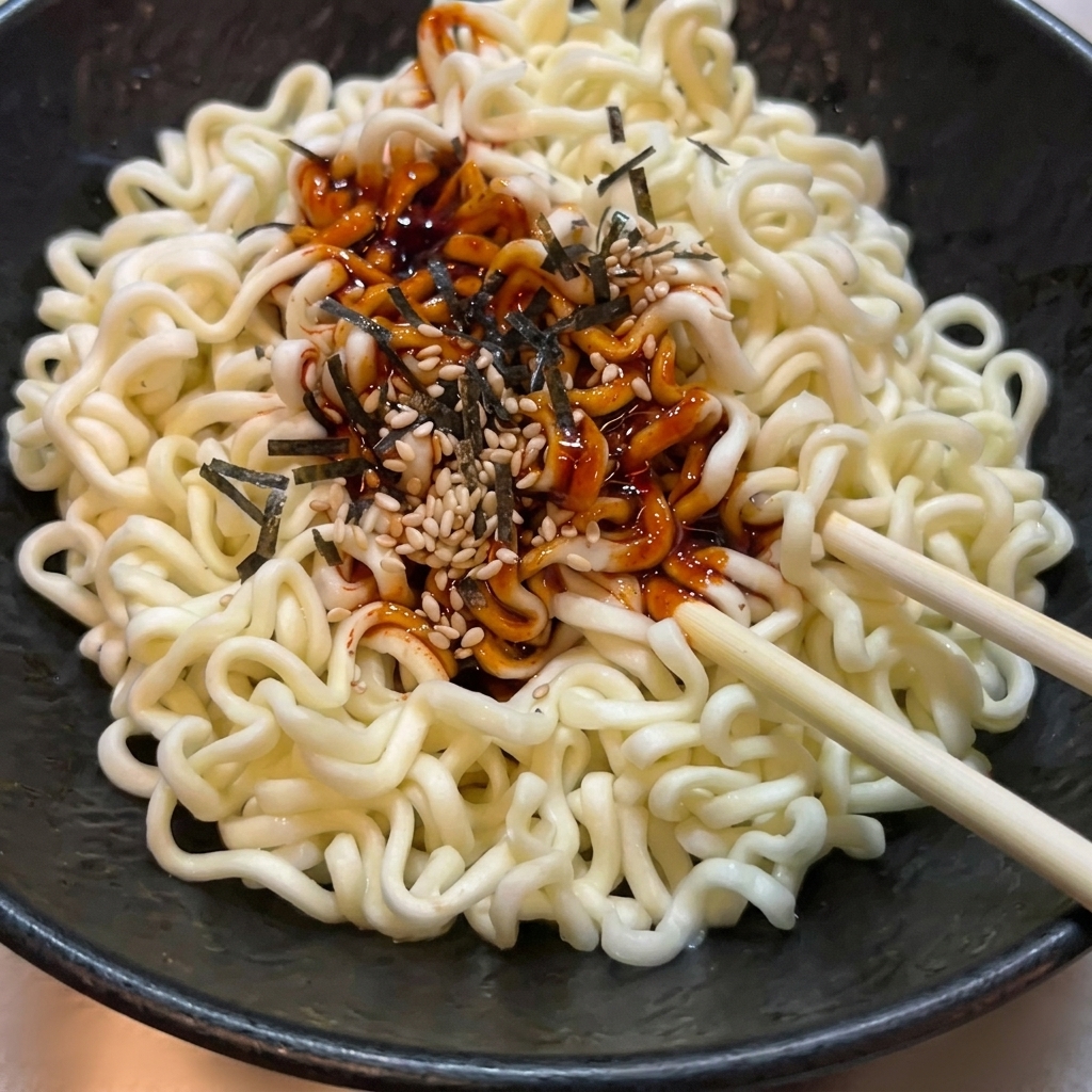 Gekochte, gewellte Ramen-Nudeln in einer schwarzen Schüssel werden mit roter scharfer Sauce, Sesam und Algen vermengt.