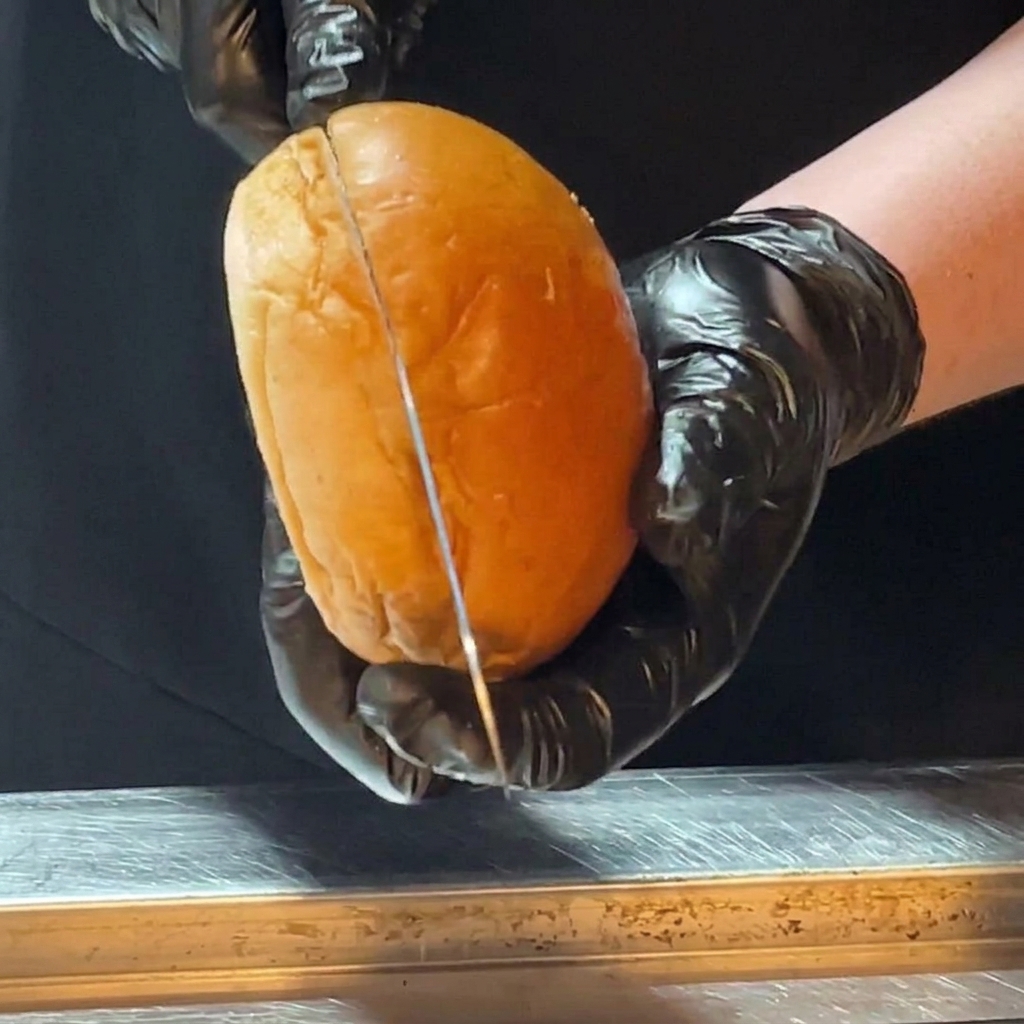 Hände in schwarzen Handschuhen halbieren ein glänzendes Brioche-Brötchen mit einem langen Messer.