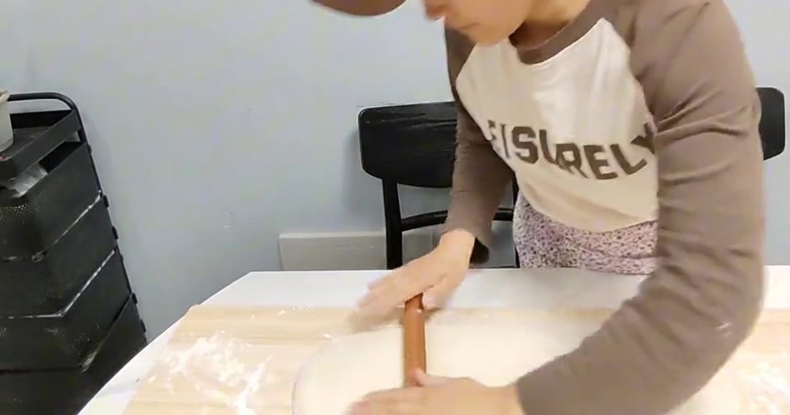 Eine Person verwendet ein hölzernes Nudelholz, um einen Teigblock auf einem bemehlten Holzbrett flach zu rollen.