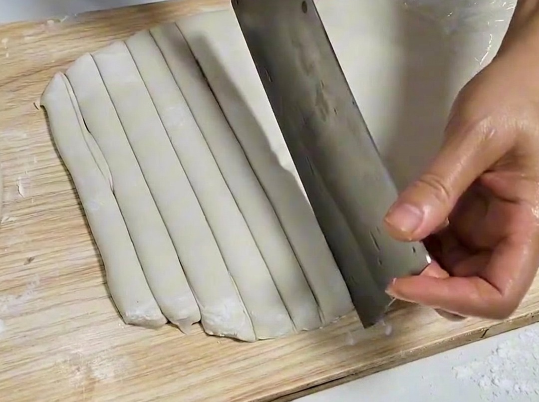 Ein Teigblatt wird mit einem Metallbeil in gleichmäßige Streifen auf einem Holzbrett geschnitten.