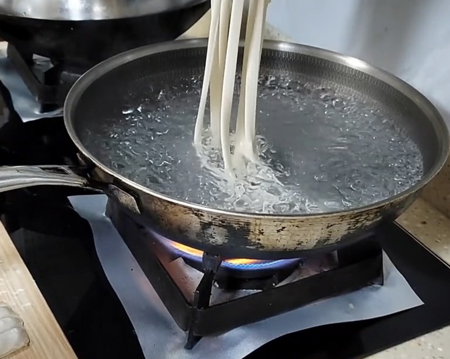 Eine Hand dehnt einen Teigstreifen zu einer langen Nudel, bevor sie in einen Topf mit kochendem Wasser gegeben wird.