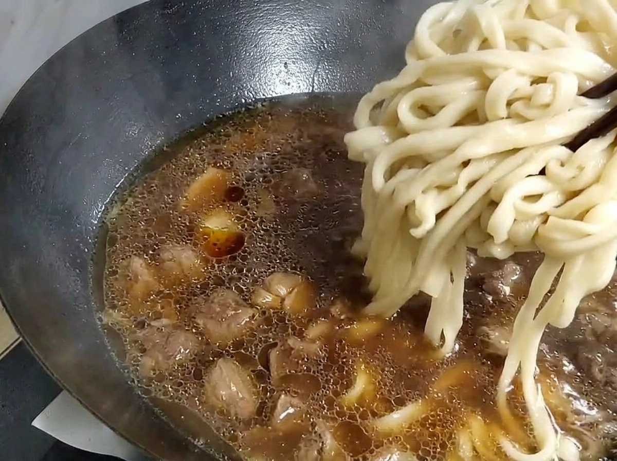 Dicke Nudeln werden mit Stäbchen aus einem Topf mit Wasser gehoben und in einen Wok mit Suppenbasis gelegt.