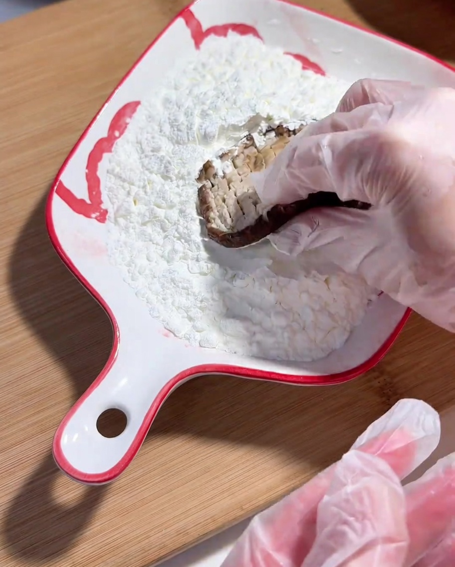 Eine Hand mit Plastikhandschuh drückt die eingeschnittene Seite eines Shiitake-Pilzes in Maisstärke.