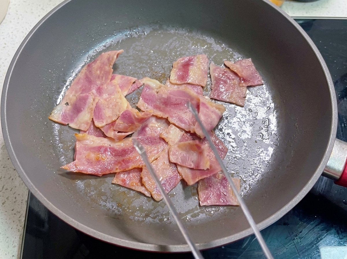 Eine Pfanne enthält gebratenen Speck, über den die verquirlte Eimischung gegossen wird.