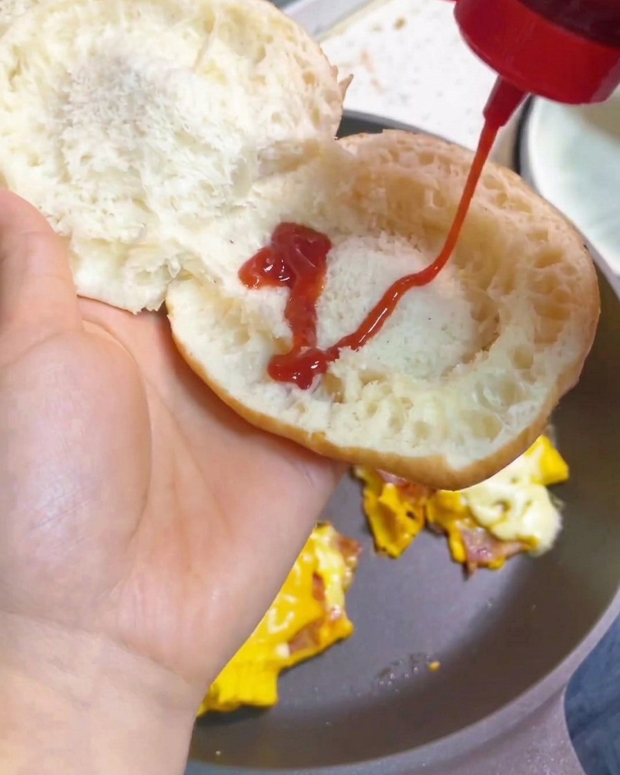 Eine Hand drückt Ketchup zickzackförmig aus einer Flasche auf die Innenseite eines getoasteten Burger-Brötchens.