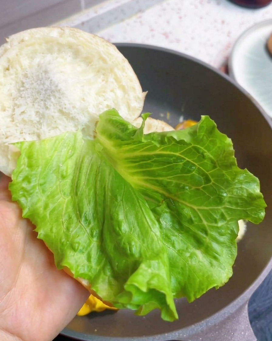 Ein Frühstücksburger wird mit frischem Salat und dem Speck-Ei-Käse-Patty in den gerösteten Brötchen zusammengesetzt.