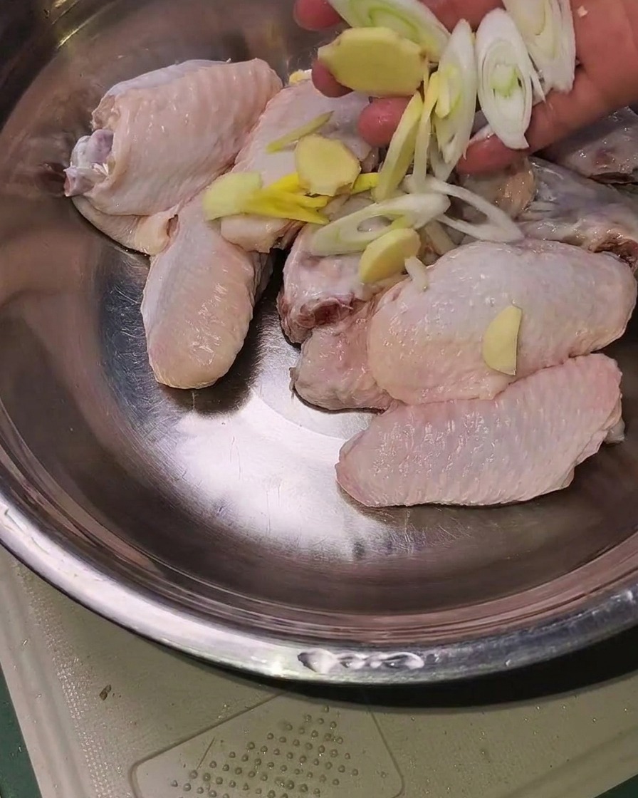 Rohe Hähnchenflügel in einer Metallschüssel, bestreut mit Ingwer, Knoblauch und Frühlingszwiebeln.