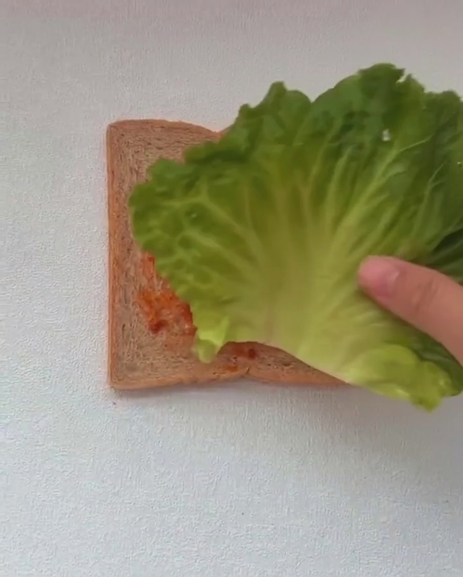 Zwei große grüne Salatblätter werden auf eine mit roter Sauce bestrichene Toastscheibe gelegt.