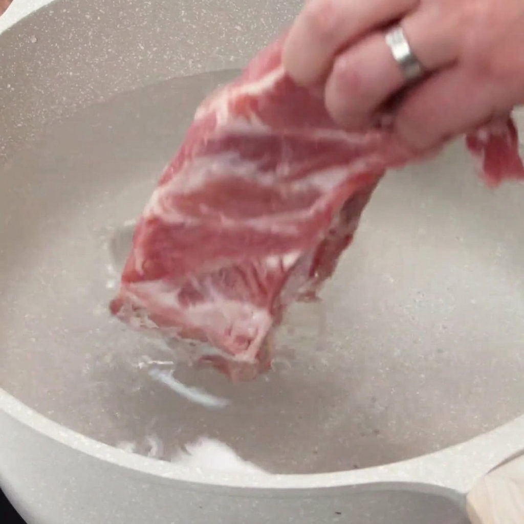 Eine Hand legt rohe Schweinerippchen in einen Topf mit kaltem Wasser.