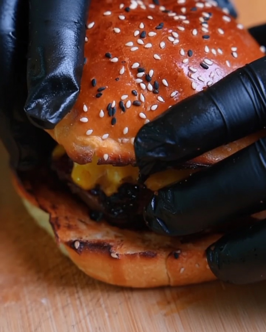 Nahaufnahme eines Burgers, der mit dem oberen Brötchen fertiggestellt wird, gehalten von behandschuhten Händen.