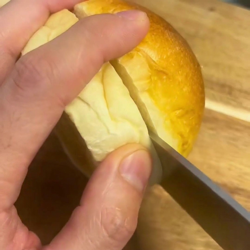 Ein Messer schneidet ein weiches Burgerbrötchen auf einem Holzschneidebrett in zwei Hälften.