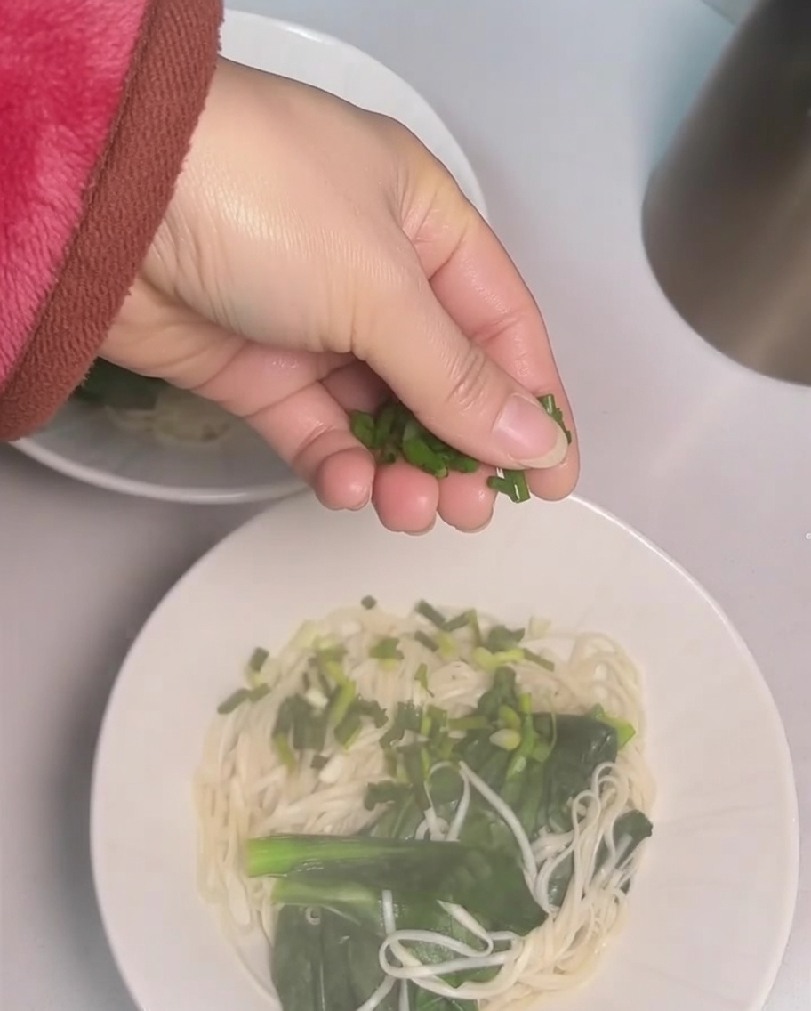 Eine Hand streut gehackte Frühlingszwiebeln über eine Schale Nudeln.