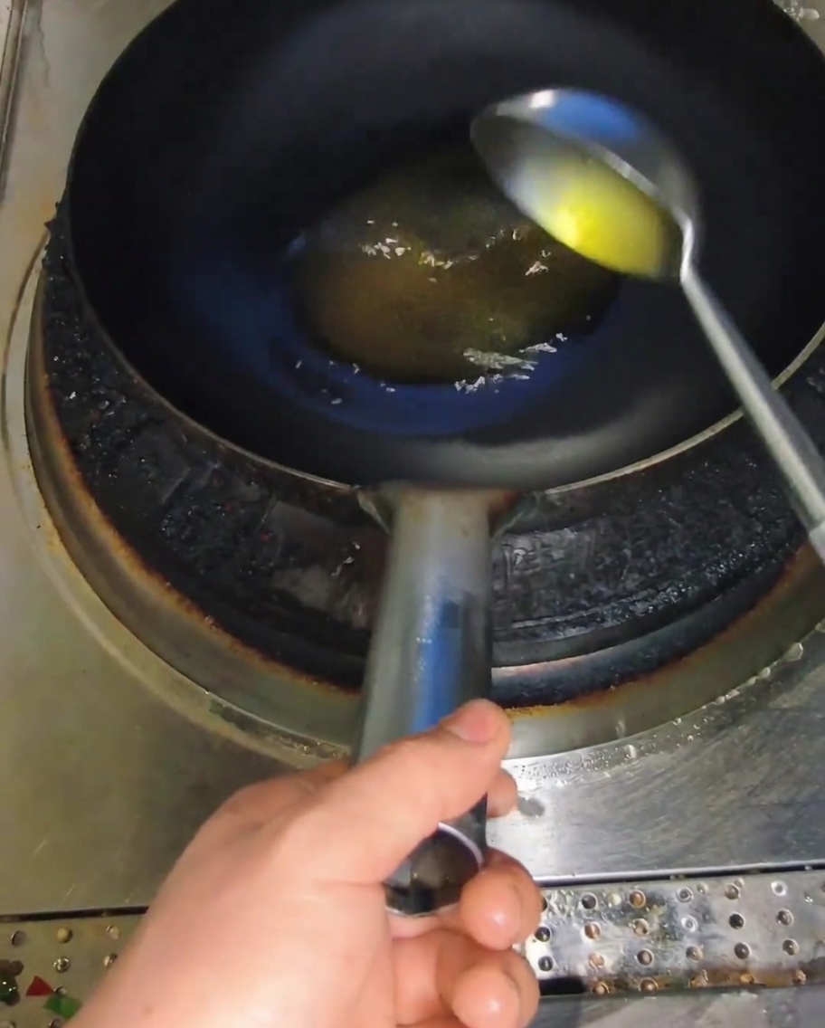 Eine Metallkelle gießt gelbes Speiseöl in einen heißen, dunklen Wok.