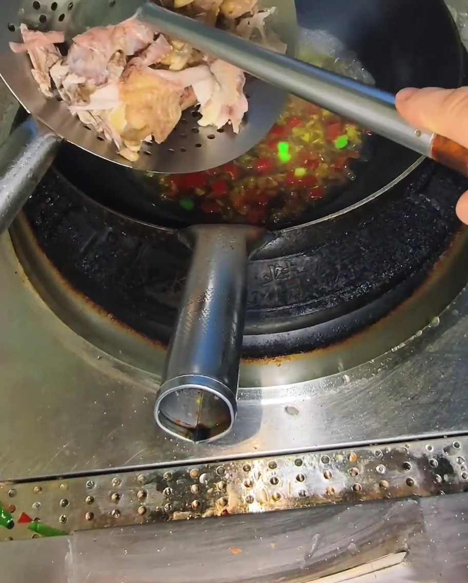 Ein Metall-Schaumlöffel gibt Stücke von zerkleinertem Hähnchen in einen Wok mit kochender, würziger Brühe.