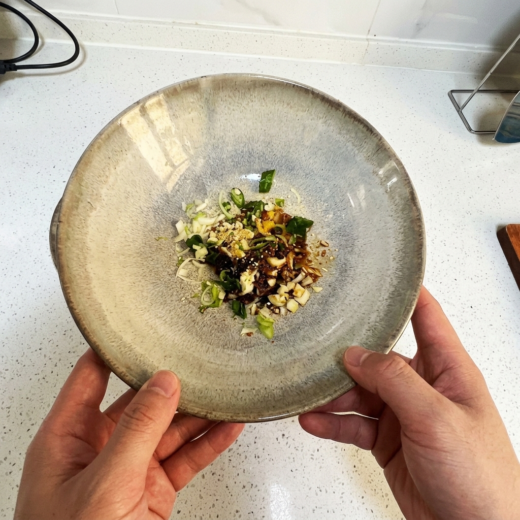 Eine von zwei Händen gehaltene Keramikschale mit gehackten Frühlingszwiebeln, gehacktem Knoblauch und flüssigen Gewürzen.