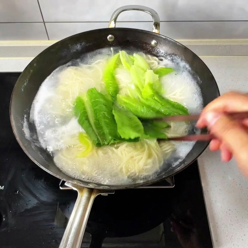Dünne Nudeln und frisches Blattgemüse kochen in einem Wok mit sprudelndem Wasser und werden mit Holzstäbchen umgerührt.