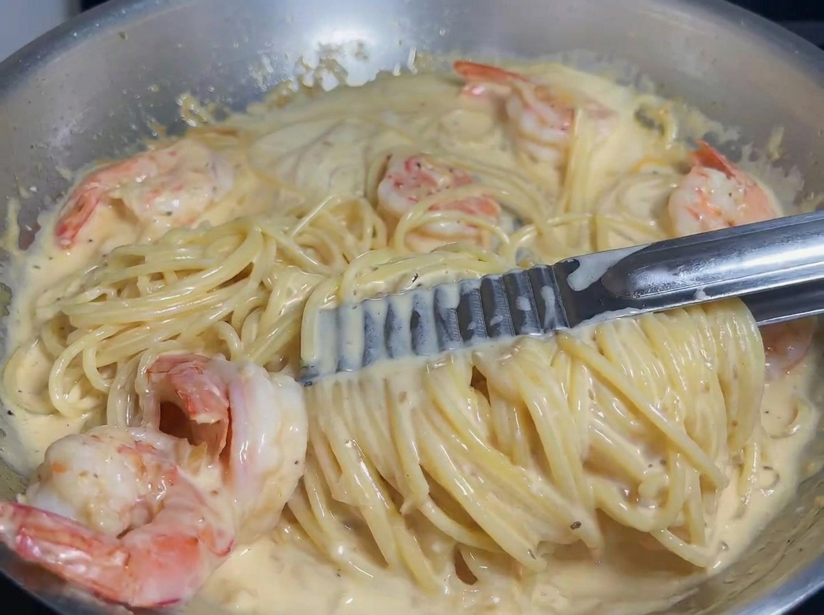 Pasta und Garnelen werden mit einer Zange in einer cremigen Sauce geschwenkt, um eine gleichmäßige Umhüllung zu gewährleisten.