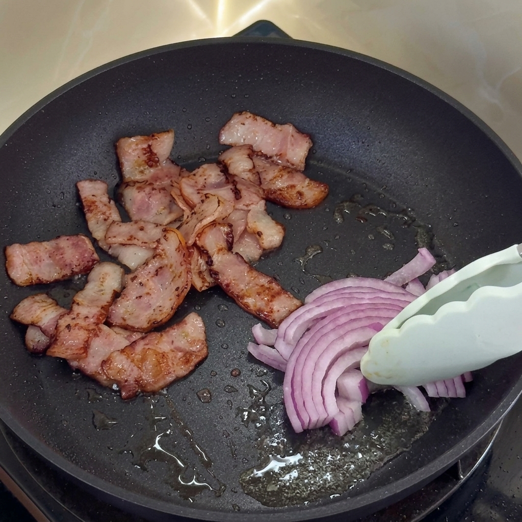 Geschnittene rote Zwiebeln garen im Speckfett neben knusprigem Speck.