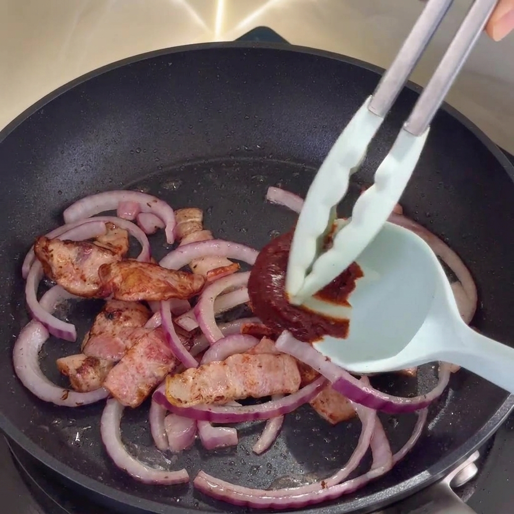 Koreanische scharfe Sauce wird unter die Speck-Zwiebel-Mischung in der Pfanne gerührt.
